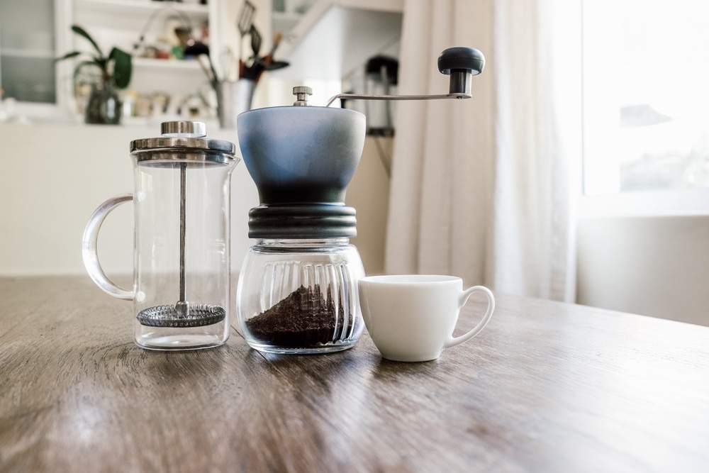 A manual coffee grinder on a wooden table next to a small saucer and French press.