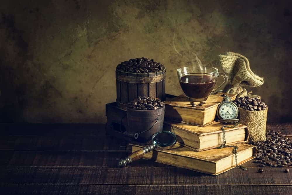A cup of coffee on top of a stack of books with a clock and buckets of coffee beans.