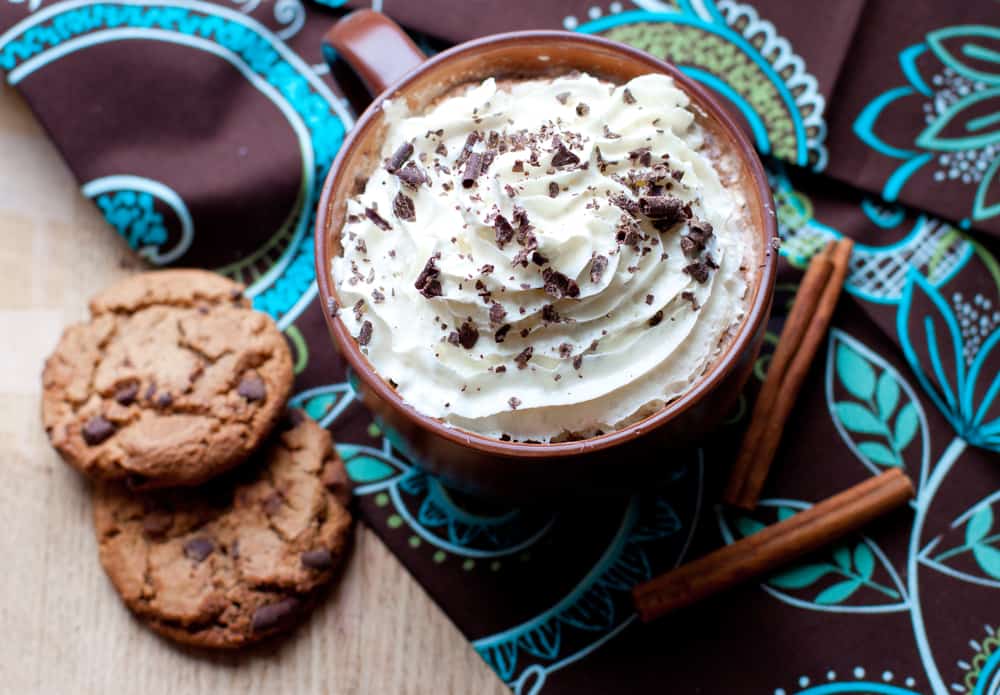 Hot chocolate with whipped cream and chocolate shavings next to chocolate chip cookies and cinnamon sticks